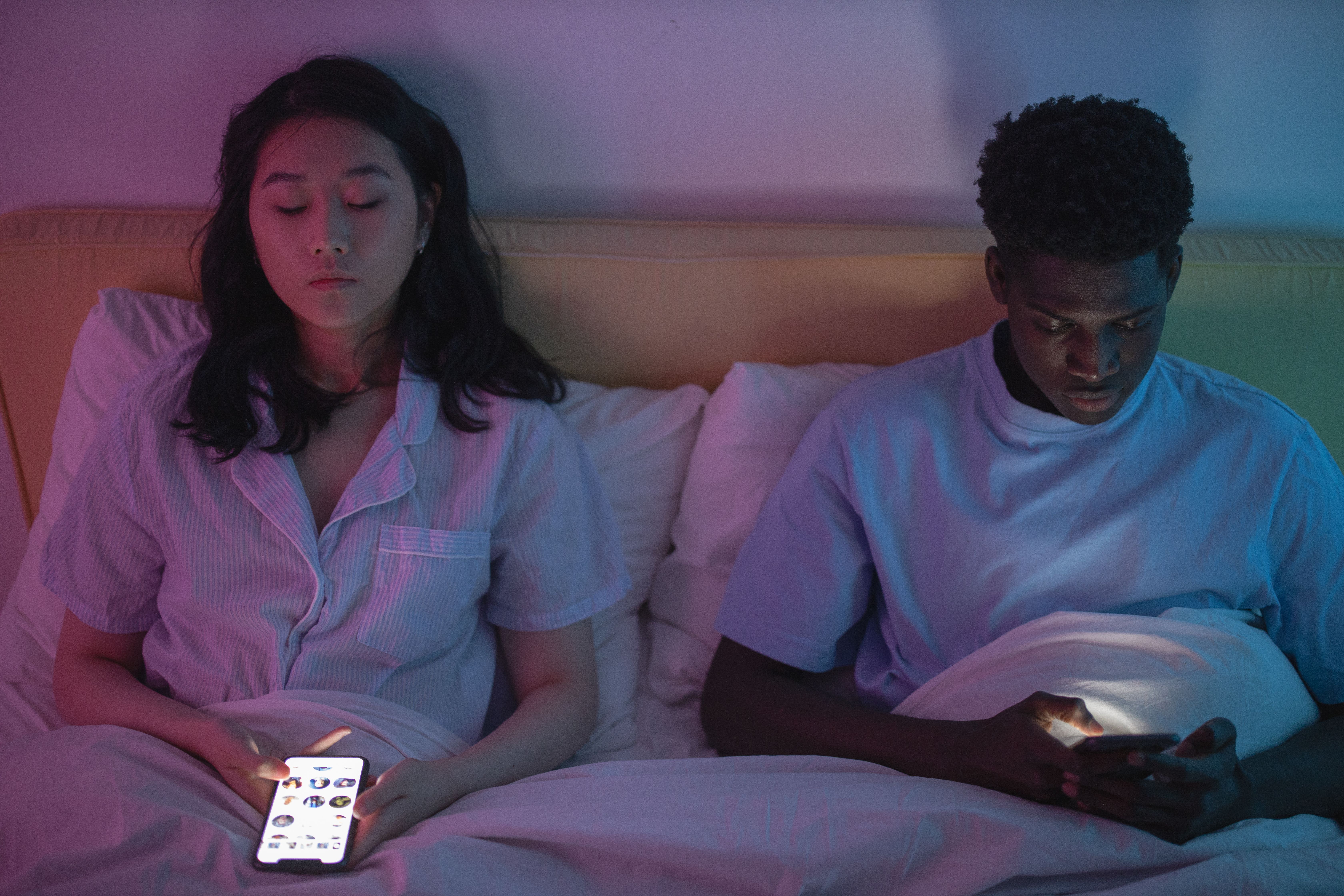 A couple lying in bed at night using separate smartphones, showing how digital devices are part of modern intimate relationships.