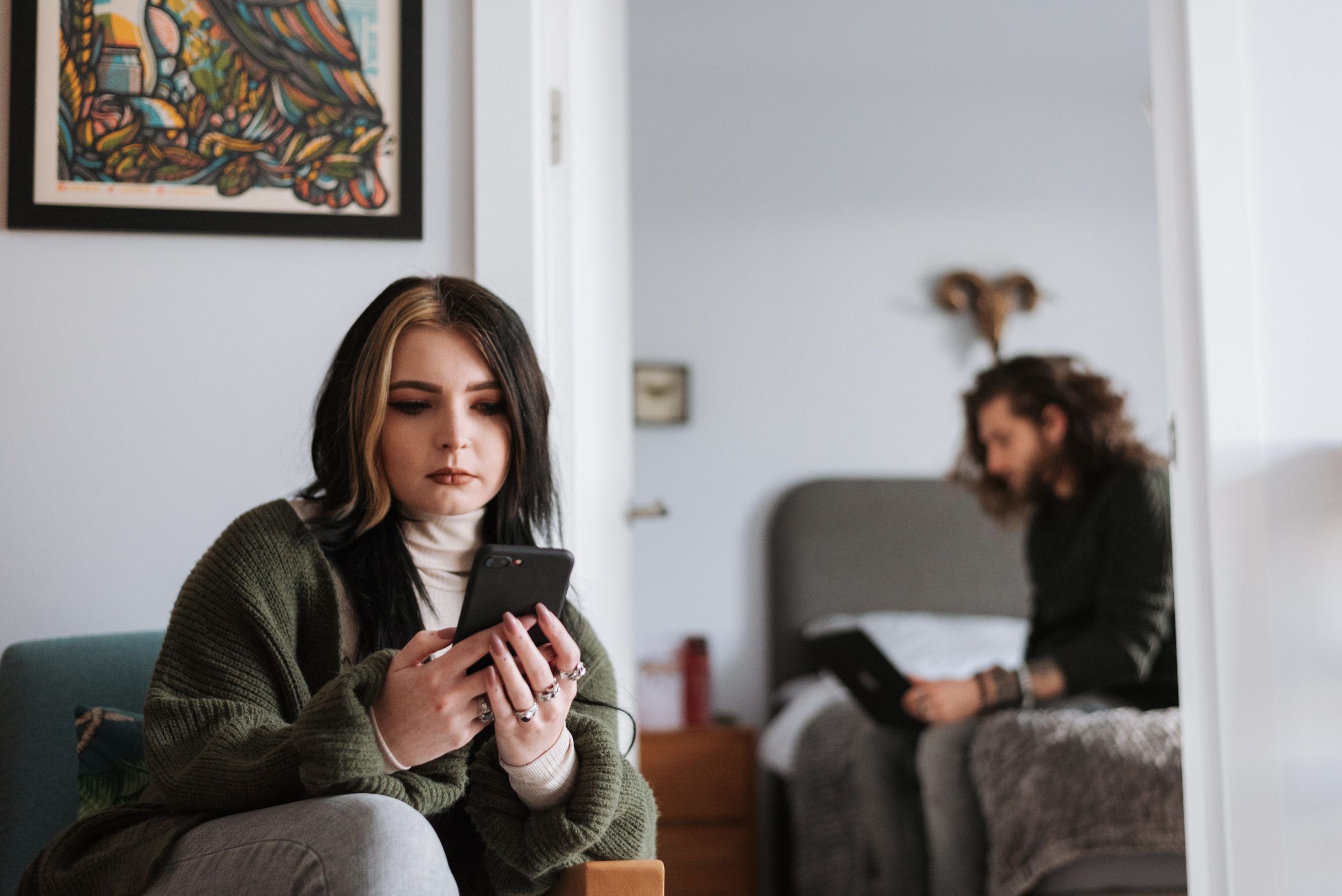 A woman messages on her phone at home while her partner works nearby on a laptop, illustrating privacy within a shared relationship.